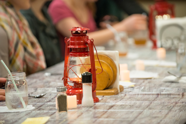 A close-up shot of the dining table decor. The table is covered by a faux wood plastic table cloth. Several red battery operated lanterns and white LED candles provide dim lighting. A wooden napkin holder stuffed with white paper napkins, salt and pepper shakers, silverware, handwritten menus, and mason jars with straws are also present on the table. 