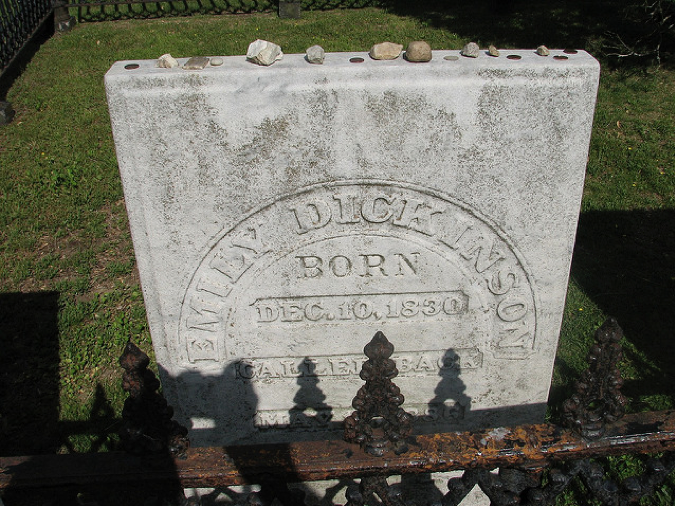 A close-up of the plot of Emily Dickinson's grave. A rectangular, gray-white tombstone is seen behind a rusty iron fence. Rocks and pennies left by Dickinson admirers sit on the flat top of the stone.