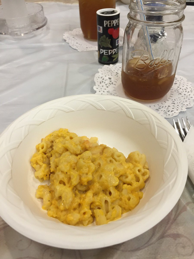A close-up shot of yellow mac and cheese in a white paper bowl on a white plastic table cloth. A fork wrapped in a white napkin, a less-than-half-full mason jar of iced tea with a straw sitting atop a white doily, and a pepper shaker can also be seen in the upper-right portion of the photo. 