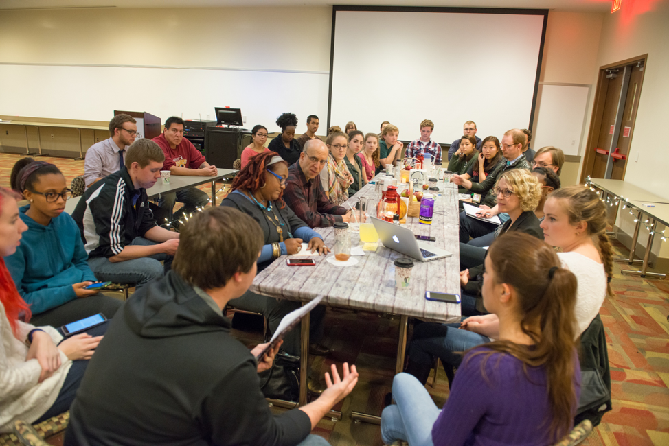 Everyone involved in the event is gathered around the dining table during the "After-Dinner Debrief." In this shot, most people are turned toward a male student who is sharing his thoughts. He is seated at the far end of the table closest to the camera.