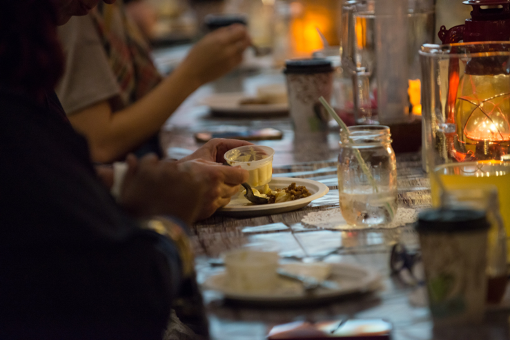 This photo shows the table during dessert. The room is dark at this point, aside from the glow of the lanterns, candles, and projected firelight. The students' faces are not shown, but the photo reveals one of their hands scooping vanilla ice cream out of a small plastic container to put on top of the apple crisp. 