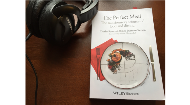 Our course text, The Perfect Meal: The Multisensory Science of Food and Dining, is pictured along with large black Koss headphones on top of a dark brown wooden table.