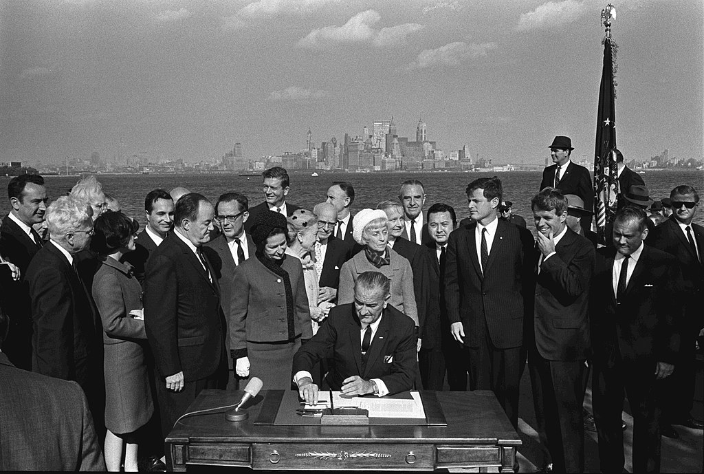 Outdoor photograph of President Lyndon B. Johnson signing an immigration bill with a crowd of onlookers surrounding him with the New York City skyline in the background.