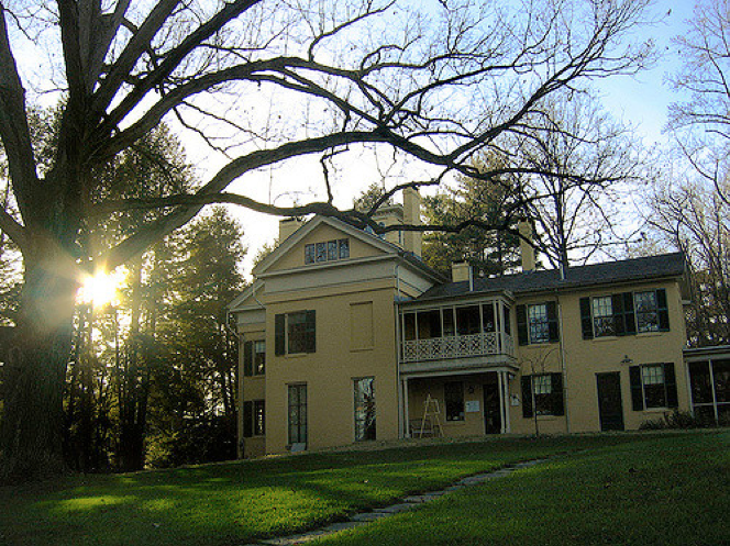 A large, yellow, Federal-style house is seen from a distance against a light blue sky. The sun is shining through pine trees behind the home and a large leafless tree sits in the expansive green front yard.