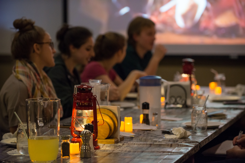 A close-up photo of the table during Sonic Palate Cleanser \#2. Four students are seen sitting and talking in the soft light from the projected fire, lanterns, and candles as they wait for the final course. 