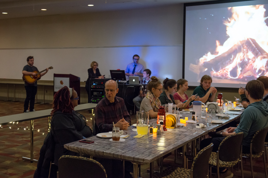 A photo captured during the tail end of the main course. The overhead lights are dimmed very low. There is a large campfire projected on to the screen at the far end of the dining table. Diners eat and chat, faces lit by the glowing lights from the fire, the lanterns and candles, and the Christmas lights that surround the table. A student playing an acoustic guitar can be seen at the far end of the room beside a computer station where Ceraso observes her two students operating the sound for the event. 